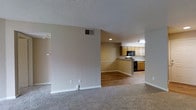 This single-floor residence offers a practical and welcoming layout designed for comfortable everyday living. The living area is finished with wall-to-wall carpet in a neutral tone, creating a soft and inviting atmosphere. Warm beige walls and white baseboards run throughout, lending the space a clean, cohesive aesthetic. A textured popcorn ceiling adds a classic touch overhead, while a ceiling-mounted HVAC vent ensures consistent climate control throughout the space.

The open layout provides a clear sightline from the living area into the kitchen, which is separated by a half-wall peninsula that doubles as a casual dining or prep surface. The kitchen transitions to hardwood-style flooring, offering a durable and easy-to-clean surface underfoot. Warm wood-toned cabinetry lines the upper and lower walls, providing ample storage. Black appliances, including a range and an over-the-range microwave, add a modern contrast to the warm cabinetry. Recessed lighting illuminates the kitchen area, complemented by an overhead light fixture. The overall ambiance is warm and functional, with a straightforward floor plan that makes efficient use of the available space.
