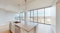 This single-floor residence is defined by its striking floor-to-ceiling glass curtain wall, which spans the living and bedroom areas to deliver expansive, unobstructed city views. The open-concept living space features wide-plank light hardwood flooring and a clean, contemporary aesthetic that maximizes the sense of openness and natural light.

The kitchen is a well-appointed centerpiece, featuring a large island with a quartz countertop, undermount sink with a high-arc faucet, and pendant lighting overhead. Soft-tone shaker cabinetry, a decorative tile backsplash, stainless steel appliances including a wall oven and French door refrigerator, and a vented range hood complete the space.

A hallway connects the living area to the private quarters, where the primary bedroom offers plush carpeting, floor-to-ceiling windows, and access to a walk-in closet with built-in shelving. The en-suite primary bathroom features a double vanity with gold-tone fixtures, tile flooring, and a glass-enclosed walk-in shower with dark tile surround. A separate powder room with pedestal sink and wood-look flooring provides guest convenience. The unit also includes a dedicated laundry room with side-by-side washer and dryer and overhead shelving.