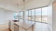 This single-floor residence is defined by its striking floor-to-ceiling glass curtain wall, which spans the living and bedroom areas to deliver expansive, unobstructed city views. The open-concept living space features wide-plank light hardwood flooring and a clean, contemporary aesthetic that maximizes the sense of openness and natural light.

The kitchen is a well-appointed centerpiece, featuring a large island with a quartz countertop, undermount sink with a high-arc faucet, and pendant lighting overhead. Soft-tone shaker cabinetry, a decorative tile backsplash, stainless steel appliances including a wall oven and French door refrigerator, and a vented range hood complete the space.

A hallway connects the living area to the private quarters, where the primary bedroom offers plush carpeting, floor-to-ceiling windows, and access to a walk-in closet with built-in shelving. The en-suite primary bathroom features a double vanity with gold-tone fixtures, tile flooring, and a glass-enclosed walk-in shower with dark tile surround. A separate powder room with pedestal sink and wood-look flooring provides guest convenience. The unit also includes a dedicated laundry room with side-by-side washer and dryer and overhead shelving.