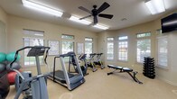 Located at 3006 E 51st St in Tulsa, Oklahoma, this fitness center is a well-appointed, single-story exercise facility designed to support a comprehensive workout experience. The space is thoughtfully organized into distinct functional zones, accommodating both cardio training and strength work within an open, cohesive layout.

The cardio area is equipped with treadmills, elliptical machines, and cycling bikes, all positioned to take advantage of the abundant natural light that streams through numerous windows fitted with slatted blinds. Transom windows positioned above standard windows enhance brightness throughout, creating an inviting and energizing atmosphere. A ceiling fan provides additional air circulation, contributing to user comfort during exercise.

A large wall mirror spans one side of the room, visually expanding the space and allowing users to monitor their form. Workout reference charts are posted along the walls, offering guidance on exercises and heart rate targets. A wall-mounted rack holds resistance bands and suspension training equipment, providing versatile strength-training options.

The strength zone features a dumbbell rack and an adjustable bench, complemented by a wall-mounted flat-screen television. Exercise balls add flexibility training options. Neutral-toned walls and light carpeted flooring contribute to a clean, functional ambiance, while views of surrounding greenery add a calming, natural element to the environment.