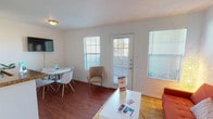 This single-floor unit offers a practical and efficiently designed layout across one bedroom and one bathroom. The living area features rich hardwood-style flooring, multiple windows, and a private exterior door that fills the space with natural light while providing direct outdoor access. A wall-mounted TV and defined dining area make the living space versatile for both relaxing and entertaining.

The kitchen is positioned adjacent to the living area and includes a peninsula with granite-style countertops that doubles as a breakfast bar. Medium-toned wood cabinetry, a black refrigerator, and a sink with ample counter space complete the functional cooking area.

The bedroom is carpeted for comfort and equipped with a ceiling fan, a wide closet with shelving, and a window for natural light. Notably, a stacked washer and dryer unit is conveniently located within the bedroom area, adding practical in-unit laundry access.

The bathroom features tile flooring, a vanity with granite-style countertops and a wood cabinet base, a large mirror, and a shower-tub combination. The overall aesthetic throughout the unit is clean and neutral, with white walls providing a bright and straightforward living environment.