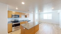 This single-floor residence is designed with a clean, modern aesthetic that prioritizes functionality and cohesion. The kitchen serves as a central feature of the space, equipped with warm honey-toned cabinetry, speckled granite countertops, and a full suite of stainless steel appliances including a French door refrigerator with ice maker, electric range, built-in microwave, and dishwasher. A granite-topped kitchen island provides additional workspace and naturally defines the transition into the open-concept living area. Recessed lighting and a textured ceiling add depth to the kitchen and living space, while light-toned hardwood flooring flows continuously throughout, lending warmth and visual consistency.

The living area benefits from natural light through windows fitted with horizontal blinds, creating a bright and airy atmosphere. A bedroom features the same hardwood flooring, a ceiling fan with light fixture, and a window offering natural light and exterior views.

The bathroom is thoughtfully appointed with a wide granite-topped vanity, dual undermount sinks, warm wood cabinetry with brushed nickel hardware, a large mirror, and a tub-shower combination. The overall design of the space is cohesive and contemporary, with neutral tones and quality finishes throughout.