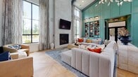 Located at 8400 Palmetto Commerce Pkwy in Ladson, South Carolina, this single-floor clubhouse delivers an impressive blend of contemporary design and inviting comfort. The space is anchored by a dramatic vaulted ceiling with exposed dark beams and white-paneled shiplap, which together create a grand sense of height and openness. A sculptural, branch-inspired chandelier hangs as the centerpiece, casting warm ambient light throughout.

The main lounge area features expansive herringbone-patterned hardwood floors that extend across the entire space, enhancing its generous proportions. Bold teal board-and-batten wall paneling forms a striking architectural backdrop, housing built-in shelving filled with curated décor and books. A floor-to-ceiling stacked stone fireplace with a mounted television anchors one wall, flanked by tall, black-framed windows dressed with flowing drapery that flood the room with natural light.

The open layout transitions seamlessly into a game area near the entry, complete with a foosball table and additional wall art installations. Black-framed glass doors lead to an outdoor area, further connecting the interior to the surrounding landscape. A dining and work zone with a dark wood table adds functional versatility. The overall ambiance is modern yet warm, combining rich tones, curated furnishings, and architectural details into a sophisticated and welcoming communal environment.