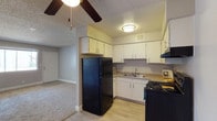 This single-floor residential space is designed for comfortable, practical living. The kitchen is a standout feature, outfitted with crisp white cabinetry that provides ample storage along both upper and lower sections. Black appliances, including a full-size refrigerator and an electric range with overhead ventilation, add a bold contrast against the light-toned countertops and wood-look vinyl flooring. A double-basin sink is neatly positioned beneath a flush-mount ceiling light, and a visible HVAC vent ensures year-round climate comfort. The kitchen layout is efficient and well-organized, making it easy to navigate during everyday use. A textured ceiling adds architectural character throughout the space. Adjacent to the kitchen, the living area transitions to plush carpeting, creating a warm and inviting atmosphere. A ceiling fan with wooden blades is centrally positioned, promoting air circulation while adding a classic touch to the room. A sliding closet door is visible along one wall, offering convenient built-in storage. Together, the spaces form a cohesive, functional layout well-suited for everyday residential living.
