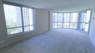 This high-rise unit offers a compelling blend of urban energy and interior comfort, defined by its striking floor-to-ceiling wraparound windows that flood the space with natural light and frame panoramic views of the surrounding city skyline. The generously proportioned main living area is finished with plush wall-to-wall carpeting in a neutral tone, creating a warm and inviting atmosphere that complements the clean, minimalist design of the space. Smooth, painted walls in a soft off-white hue enhance the bright, airy feel, while the curved window bay adds an architectural touch that sets this space apart from a standard rectilinear layout. The elevated position of the unit is immediately apparent through the windows, where neighboring high-rise towers and city rooftops stretch into the distance, offering a dynamic urban backdrop throughout the day. The open layout provides excellent flexibility for furniture arrangement and living configurations. The overall ambiance is one of spaciousness, brightness, and modern urban living, making it well-suited for those who appreciate natural light, expansive views, and a clean contemporary aesthetic.