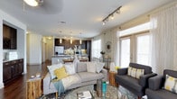 This single-floor residence offers a thoughtfully designed open-concept layout that connects the living area and kitchen in one fluid, welcoming space. The living room is anchored by a comfortable seating arrangement, complemented by a patterned area rug and a glass-topped coffee table, creating an inviting atmosphere for relaxing or entertaining. Large windows dressed with sheer white curtains flood the space with abundant natural light, while a glass door provides direct access to the outdoors. Track lighting along the ceiling adds a contemporary touch and ensures the space is well-illuminated throughout. The kitchen is a standout feature, showcasing rich dark cabinetry, a stainless steel refrigerator, and a central island with pendant lighting overhead — ideal for casual dining or meal preparation. Light-toned countertops contrast beautifully with the darker cabinetry, lending the kitchen a refined, balanced aesthetic. Hardwood flooring flows throughout, tying the spaces together cohesively. The overall ambiance is modern yet warm, with a neutral color palette accented by soft yellows and blues that add character and vibrancy without overwhelming the senses. This well-appointed space is designed for comfortable, stylish everyday living.
