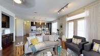 This single-floor residence offers a thoughtfully designed open-concept layout that connects the living area and kitchen in one fluid, welcoming space. The living room is anchored by a comfortable seating arrangement, complemented by a patterned area rug and a glass-topped coffee table, creating an inviting atmosphere for relaxing or entertaining. Large windows dressed with sheer white curtains flood the space with abundant natural light, while a glass door provides direct access to the outdoors. Track lighting along the ceiling adds a contemporary touch and ensures the space is well-illuminated throughout. The kitchen is a standout feature, showcasing rich dark cabinetry, a stainless steel refrigerator, and a central island with pendant lighting overhead — ideal for casual dining or meal preparation. Light-toned countertops contrast beautifully with the darker cabinetry, lending the kitchen a refined, balanced aesthetic. Hardwood flooring flows throughout, tying the spaces together cohesively. The overall ambiance is modern yet warm, with a neutral color palette accented by soft yellows and blues that add character and vibrancy without overwhelming the senses. This well-appointed space is designed for comfortable, stylish everyday living.