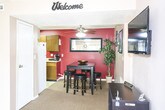 This single-floor commercial space is thoughtfully arranged to accommodate both client-facing and operational needs. The reception area greets visitors with neutral-toned walls, carpeted flooring that transitions to tile near the entrance, and a pair of dark upholstered chairs arranged around a small side table. Decorative diamond-shaped wall art adds a touch of personality, while a wall-mounted mailbox and framed informational signage contribute to the professional atmosphere. A sliding glass door floods the space with natural light and offers a street-level exterior view, complemented by a ceiling fan overhead.

Adjacent to the reception area, a dining or break space features a bold red accent wall, a ceiling fan with light fixture, a raised dark wood table with bar stools, and a wall-mounted mirror. The adjoining kitchen is equipped with wood-tone cabinetry, a white refrigerator, and a countertop with a coffee maker, providing practical functionality.

A carpeted hallway extends further into the space, lined with framed certifications and photographs, and leads to multiple private rooms or offices accessible through individual doors. A textured ceiling and floor vent are consistent throughout, giving the space a cohesive, utilitarian character.