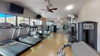 This fitness center offers a comprehensive workout environment designed to accommodate a wide range of exercise routines. The space features durable, speckled rubberized flooring throughout, providing a practical and easy-to-maintain surface suited for both cardio and strength training activities.

The cardio section is outfitted with multiple treadmills, elliptical trainers, and a recumbent bike, all from True Fitness, arranged along one side of the room. Wall-mounted flat-screen televisions are positioned for viewing during workouts. A ceiling fan overhead aids in air circulation, complemented by wall-mounted fans for additional ventilation.

Floor-to-ceiling mirrors line one wall, visually expanding the space and allowing users to monitor their form. The strength training area includes a cable machine, weight stack machines for targeted muscle groups, and a freestanding dumbbell rack.

Near the entrance, a bulletin board displays facility rules and community notices alongside a water cooler and waste bin for convenience. A glass-panel door opens to the exterior, allowing natural light to filter into the space. Soft wall sconces provide ambient lighting, contributing to a clean, functional atmosphere throughout.