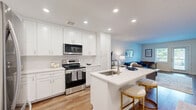 Located at 138 Mt Auburn Ave in Auburn, Maine, this thoughtfully designed single-floor residence offers a clean, modern aesthetic across a well-utilized layout. The open-concept kitchen and living area serves as the heart of the home, where crisp white shaker cabinetry extends to the ceiling, providing generous storage. Stainless steel appliances—including a range, microwave, and refrigerator—complement the bright white countertops. A center island with an undermount sink and seating for two creates a natural gathering space that flows seamlessly into the living area. Warm hardwood-style flooring runs throughout the main living space, adding a welcoming contrast to the bright white kitchen. The living room is generously proportioned, filled with natural light from windows and a glass-panel exterior door, creating an open and airy ambiance. A hallway leads to the private bedroom, which features plush carpet flooring, a natural light-admitting window with blinds, and a dedicated closet for ample storage. The bedroom's neutral palette and soft finishes contribute to a calm, restful atmosphere. Recessed lighting throughout the unit ensures consistent, modern illumination. This residence combines functional design with contemporary finishes for comfortable everyday living.