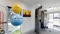 This fitness facility offers a well-appointed workout environment designed to support a wide range of exercise routines. The space is finished with clean white walls, recessed lighting, and carpeted flooring, creating a bright and motivating atmosphere conducive to physical activity.

A multi-functional cable machine takes a prominent position in the room, providing users with versatile resistance training options. Complementing the strength equipment is a colorful collection of stability balls in varying sizes, adding a playful yet functional element ideal for balance, core, and flexibility training.

Natural light filters in through windows that overlook lush greenery, offering an inviting outdoor connection that enhances the overall ambiance. Inspirational fitness artwork adorns the walls, including a striking canvas print depicting silhouetted figures in yoga poses against a vivid sunset, reinforcing the space's focus on health and wellness.

The layout is thoughtfully organized to maximize usability while maintaining an open, uncluttered feel. The combination of natural light, modern equipment, and motivating decor makes this fitness space both functional and an enjoyable environment for users seeking a comprehensive workout experience.