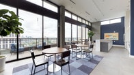This contemporary communal space makes a striking impression with its soaring floor-to-ceiling windows that span the entire exterior wall, flooding the interior with natural light and offering expansive panoramic views of the surrounding cityscape. The elevated position of the space enhances the sense of openness, creating an airy and sophisticated atmosphere throughout.

The layout features a flexible seating arrangement with multiple round dining tables paired with sleek, modern chairs, all set atop a geometric patchwork area rug in muted tones of grey and blue. The combination of light tile flooring and the soft rug delineates the social zones seamlessly. Pendant lighting and a ceiling-mounted track lighting system provide warm, focused illumination to complement the abundant daylight.

Architectural details include exposed concrete columns and walls, which lend an industrial character balanced by refined finishes and carefully selected furnishings. A lush potted plant adds a natural accent, while an adjacent area features cabinetry finished in deep navy blue, suggesting an informal meeting or refreshment zone. Beyond the windows, a generous outdoor terrace with railing is visible, extending the usable social space further. The overall ambiance is modern, professional, and inviting — well-suited for collaborative gatherings or casual meetings.