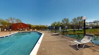 This outdoor pool facility in Evansville, Indiana, offers a generously sized rectangular swimming pool with clear blue water, framed by a broad concrete deck that provides ample space for relaxation and circulation. The pool features built-in metal entry ladders at multiple points and clearly marked depth indicators along its edges, ensuring safe and informed use by swimmers of all levels. Posted warning signage notes that there is no lifeguard on duty and that children under 14 must be accompanied by a responsible adult.

Surrounding the pool deck, numerous white lounge chairs are arranged to take full advantage of the open sky and abundant natural sunlight. Globe-style lamp posts are positioned throughout the area, providing illumination for evening use. The entire pool area is enclosed by a chain-link fence, offering both security and visibility to the surrounding grounds, which include mature trees and open green space.

Adjacent to the pool, a brick building with a pitched shingle roof serves as a supporting facility structure. A wooden exterior staircase leads to an upper-level deck on this building, adding a functional architectural element to the space. A second red brick structure sits nearby, contributing to the overall campus-like setting. The combination of open sky, lush surrounding trees, and well-maintained amenities creates a practical and inviting outdoor recreational environment.