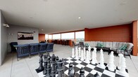 Located in the Short North neighborhood of Columbus, Ohio, this rooftop amenity deck offers an elevated communal experience with sweeping views of the city skyline. The open-air terrace is defined by large floor-to-ceiling wraparound windows and open railings that frame panoramic vistas, filling the space with abundant natural light throughout the day.

The deck is divided into several distinct activity zones. A standout feature is the oversized chess board, complete with life-size playing pieces, serving as a bold centerpiece for social interaction. Nearby, a giant Connect Four game and turf mat add to the playful atmosphere. Multiple seating areas are distributed across the terrace, including a curved sectional sofa with accent pillows, cushioned lounge chairs in both navy and neutral tones, and a wall-mounted TV for outdoor viewing.

The dining and bar area lines the windowed perimeter, furnished with tall bistro tables and bar-height chairs, ideal for gathering with views of the Columbus skyline as a backdrop. A dedicated grilling station features multiple stainless steel grills, including kamado-style ceramic cookers, perfect for outdoor entertaining.

Warm terracotta-hued accent walls contrast with the clean light-grey tile flooring and neutral paneled walls, creating a modern yet inviting aesthetic throughout this versatile rooftop retreat.