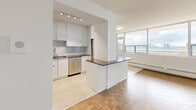 This thoughtfully designed 1-bedroom, 1-bathroom unit spans a single floor and offers a bright, open layout enhanced by classic parquet wood flooring throughout the main living areas. The open-concept kitchen and living room serve as the heart of the unit, where large floor-to-ceiling windows flood the space with natural light and frame impressive panoramic views of the city skyline and waterway beyond. The kitchen is outfitted with crisp white cabinetry, a subway tile backsplash, a dark countertop peninsula, and a full suite of stainless steel appliances including a refrigerator, dishwasher, microwave, and range — all set on clean tile flooring. The spacious living area flows seamlessly from the kitchen, offering generous room for entertaining or relaxing while taking in the elevated views. The bedroom is a well-proportioned retreat featuring a window with city views, parquet floors, and a mirrored sliding closet that adds depth and storage. The bathroom is elegantly finished with full-height white subway tile surrounding a soaking tub and shower combination, complemented by chrome fixtures and a large mirror. A connecting hallway ties the rooms together in a practical and cohesive layout.