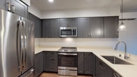 This thoughtfully designed single-floor residence showcases a clean, contemporary aesthetic carried consistently through every room. The kitchen is a standout feature, offering rich charcoal-gray shaker-style cabinetry — both upper and lower — paired with light quartz countertops and a full suite of stainless steel appliances, including a range, over-the-range microwave, refrigerator, and dishwasher. A kitchen island with pendant lighting provides additional counter space and a natural transition into the open living area. Warm wood-look plank flooring flows seamlessly throughout the main living spaces, lending a cohesive and inviting feel. The living area is generously proportioned and benefits from a sliding glass door that fills the room with natural light and provides access to an outdoor view of surrounding greenery. The bedroom is a well-sized, quiet retreat with neutral walls and the same warm flooring. The full bathroom is efficiently laid out, featuring charcoal gray cabinetry matching the kitchen, a light quartz vanity countertop with an undermount sink, a large mirror, and a walk-in shower with grab bars. A stacked in-unit washer and dryer adds notable convenience. The overall ambiance is modern, neutral, and move-in ready.