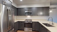 This thoughtfully designed single-floor residence showcases a clean, contemporary aesthetic carried consistently through every room. The kitchen is a standout feature, offering rich charcoal-gray shaker-style cabinetry — both upper and lower — paired with light quartz countertops and a full suite of stainless steel appliances, including a range, over-the-range microwave, refrigerator, and dishwasher. A kitchen island with pendant lighting provides additional counter space and a natural transition into the open living area. Warm wood-look plank flooring flows seamlessly throughout the main living spaces, lending a cohesive and inviting feel. The living area is generously proportioned and benefits from a sliding glass door that fills the room with natural light and provides access to an outdoor view of surrounding greenery. The bedroom is a well-sized, quiet retreat with neutral walls and the same warm flooring. The full bathroom is efficiently laid out, featuring charcoal gray cabinetry matching the kitchen, a light quartz vanity countertop with an undermount sink, a large mirror, and a walk-in shower with grab bars. A stacked in-unit washer and dryer adds notable convenience. The overall ambiance is modern, neutral, and move-in ready.