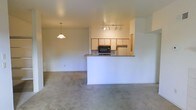 This single-floor residence offers an open-concept layout that connects the kitchen, dining, and living areas in a cohesive, functional arrangement. The kitchen is anchored by a peninsula with a granite-style countertop that serves as both a workspace and a breakfast bar, providing natural separation from the living area without sacrificing the open feel. White upper cabinetry and wood-toned lower cabinets line the kitchen walls, complemented by a built-in microwave and a refrigerator. Track lighting installed on the kitchen ceiling ensures the workspace is well-lit, while a pendant light hangs over the dining area, adding a warm focal point. The flooring transitions from the hard surface of the kitchen into soft, neutral-toned carpet that extends throughout the living and dining spaces, lending a comfortable and understated aesthetic. The walls are finished in a clean, light neutral tone throughout, contributing to a bright and airy atmosphere. The overall layout is practical and straightforward, making efficient use of the available space while maintaining an open, connected feel between the primary living areas.