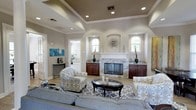 This single-story home offers a refined and welcoming atmosphere, anchored by a beautifully appointed living room that serves as the heart of the space. The room features a striking stone-clad fireplace with a classic white mantel, flanked by rich dark wood built-in cabinets that provide both style and storage. Arched wall niches frame the fireplace symmetrically, adding architectural charm and depth to the space.

The tray ceiling with crown molding adds a sense of height and elegance, while recessed lighting ensures the room is well-lit throughout. Large windows allow natural light to pour in, brightening the neutral warm-gray walls and light hardwood flooring. The open layout comfortably accommodates a generous seating arrangement, including patterned accent chairs, a plush sofa, and a glass-topped oval coffee table set atop a geometric area rug.

Tasteful decorative accents, including teal ceramic vases and wall art, introduce touches of color against the neutral palette. A hallway visible from the living area hints at additional rooms beyond, suggesting a thoughtfully connected floor plan. Overall, the space exudes a polished yet comfortable character, blending traditional architectural elements with contemporary interior styling.