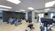 This single-level fitness room is designed to support a variety of cardiovascular workouts in a practical and well-organized setting. The space features light wood-tone flooring that lends a clean, modern aesthetic throughout. Overhead, a drop ceiling with recessed fluorescent panel lighting ensures the room is evenly and brightly illuminated, creating an energizing workout environment.

The room is outfitted with several cardio machines, including stationary bikes, an elliptical trainer, and a treadmill, providing users with multiple exercise options. Full-length mirrors line two walls, visually expanding the space and allowing users to monitor their form during workouts. The walls are painted in a two-tone scheme, with a dark charcoal accent wall contrasting against softer grey tones, contributing to a focused and modern atmosphere.

A sliding glass door or large window on one side allows natural light to filter into the room, adding to the overall brightness. Additional seating is available along the mirrored wall, and a small informational panel is mounted near the entry door. The space is functional, efficiently laid out, and well-suited for regular fitness use.