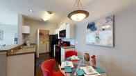 Located at 2120 Village Park Rd in Plant City, Florida, this single-story residence offers a well-designed layout with functional living spaces throughout. The kitchen is a standout feature, equipped with white cabinetry, subway tile backsplash, black appliances including a refrigerator, range, and microwave, along with a double sink and generous counter space. A breakfast bar peninsula connects the kitchen to the dining area, which comfortably accommodates a round glass dining table. An in-unit laundry area is conveniently accessible directly from the kitchen. The living room is bright and inviting, featuring large sliding glass doors dressed with vertical blinds that provide views of the lush landscaping outside, allowing abundant natural light to fill the room. The unit includes multiple bedrooms, each fitted with plush carpeting, ceiling fans, and large windows with blinds and curtain panels. One bedroom features a bold teal accent wall, while another offers warm wood-toned furnishings. Two full bathrooms are finished with wood-look flooring, white vanity cabinetry, stone-look countertops, and tub/shower combinations. Rich hardwood-style flooring runs through the kitchen and bathrooms, complementing the neutral gray tones found throughout the home, creating a cohesive and modern aesthetic.