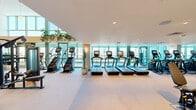 This well-appointed fitness studio in Phoenix, AZ is spread across two floors, delivering a spacious and fully equipped workout environment designed to support a wide range of training styles. The main floor serves as the primary exercise area and features soaring double-height ceilings that create an open, energizing atmosphere. Floor-to-ceiling glass windows and doors flood the space with natural light while offering panoramic views of the Phoenix skyline, adding a striking visual element to every workout. This level is comprehensively outfitted with professional-grade strength training equipment, including multi-functional cable machines, power racks, adjustable benches, and an extensive dumbbell rack with full-wall mirrors that enhance the sense of space. Cardio machines, including treadmills and stair climbers, are also present, accompanied by tasteful indoor plants that add warmth to the modern interior. A wood-railed staircase connects to the upper floor, which houses the secondary exercise room. This level features a more intimate layout with a curated selection of strength and cardio machines, recessed lighting, and large windows offering continued city views. A water fountain and storage space are conveniently available on the main level. The neutral carpeted flooring, clean white walls, and warm accent tones throughout both floors create a professional yet welcoming environment.