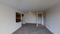 This single-floor unit is organized around a modest but functional open-plan layout. The living area is finished with wall-to-wall carpet in a neutral gray tone, providing a soft underfoot feel throughout the main space. White walls and a flush-mounted ceiling light keep the interior bright and straightforward.

A pass-through opening connects the living area to the kitchen, which features dark wood cabinetry, a dark laminate countertop that extends to form a small bar-style ledge, and a white refrigerator visible in the background. This design allows for easy interaction between the kitchen and living space while maintaining a degree of separation.

Near the front entry door, a built-in closet with a hanging rod offers convenient storage for coats and everyday items. The entryway transitions from tile flooring to the carpeted living area, clearly delineating the arrival zone from the main living space. The overall ambiance is clean and understated, with a practical layout suited for everyday living. The unit's simple, unadorned design leaves room for personalization through furnishings and décor.