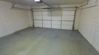 This single-level garage offers a practical and functional space designed to accommodate two vehicles with ease. The centerpiece of the space is a wide, paneled sectional roll-up door that spans nearly the full width of the garage, providing convenient access and allowing natural light to filter in when needed. A ceiling-mounted automatic garage door opener is installed directly above, adding a layer of everyday convenience. The floor is finished in smooth, durable concrete, offering a solid and easy-to-maintain surface suited for vehicle parking, storage, or workshop use. The walls are finished in a light neutral tone, keeping the interior bright and clean. The overall layout is open and unobstructed, maximizing usable floor space for vehicles, equipment, or additional storage solutions. The garage's straightforward design prioritizes utility, making it a versatile and well-appointed space for a variety of practical needs.