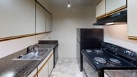 This single-floor residential unit offers a practical and well-organized layout across several distinct living areas. The spacious living room is finished with neutral-toned wall-to-wall carpeting and features a ceiling fan with integrated lighting. A large sliding glass door floods the room with natural light while providing access to an outdoor area. The open sightline connects the living room to the kitchen through a half-wall pass-through, creating a sense of continuity throughout the common areas. The galley-style kitchen is equipped with black appliances, including a coil-burner electric range, refrigerator, and dishwasher. Dark granite-style countertops complement the upper and lower cabinetry, and a single-basin stainless steel sink sits beneath a pendant light. The tile flooring adds a clean, practical finish to the kitchen space. The unit includes multiple carpeted bedrooms, each outfitted with a ceiling fan and ample closet space. One bedroom features a wide reach-in closet with a full-width hanging rod, while another benefits from a window with natural light and outdoor views. The bathroom includes a vanity with a granite-style countertop, a large wall mirror, and a separate water closet area with a bathtub, toilet, and grab bar, finished with white tile flooring throughout.