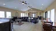 This single-floor space offers an open, airy layout defined by striking vaulted ceilings that soar overhead, fitted with multiple ceiling fans to ensure comfort throughout. The main gathering area is anchored by a generous seating arrangement featuring rich, dark leather sofas and armchairs positioned around decorative coffee tables atop a patterned area rug — creating an inviting lounge atmosphere. Large windows along the walls allow abundant natural light to fill the interior, complementing the warm, neutral-toned walls and light tile flooring that extends throughout. Recessed lighting provides additional illumination, enhancing the bright and open ambiance. Tasteful wall décor, including a large statement clock and geometric accent pieces, add personality to the space. Toward the rear, a dining area is visible with tables and chairs, suggesting the space accommodates both relaxed lounging and more formal gatherings. French doors at the back open to an outdoor area, further enhancing the sense of openness and connectivity to the exterior. Overall, this well-appointed space balances functionality and comfort, making it well-suited for socializing and community activities.