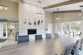 Located at 4907 Coverlane Dr in Ypsilanti, Michigan, this single-story community facility offers a well-appointed and thoughtfully designed space across two distinct areas. The centerpiece of the space is a striking conference and gathering room characterized by soaring vaulted ceilings accented with exposed wood beams, and a dramatic floor-to-ceiling stone fireplace that serves as an eye-catching focal point. Pendant lighting with glass fixtures illuminates a large conference table surrounded by sleek, modern chairs, creating an ideal environment for meetings or community events. A wall-mounted flat-screen TV provides added functionality. Light wood-look flooring and a blue abstract area rug complement the neutral warm-toned walls, while multiple large windows and French doors flood the room with natural light and offer views of the surrounding outdoor greenery. The second area of the space houses a practical on-site laundry facility, equipped with a row of front-loading washers and dryers, finished with the same clean, light flooring and neutral wall tones for a cohesive aesthetic. Together, these two spaces deliver a balance of comfort, functionality, and modern design for everyday community use.