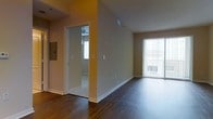 This single-floor residence offers a clean, open layout designed for comfortable everyday living. The main living area is anchored by rich, dark hardwood flooring that adds warmth and elegance throughout the space. A large sliding glass door with vertical blinds floods the room with abundant natural light while providing direct access to an outdoor balcony area, offering pleasant exterior views.

The open floor plan allows for flexible furniture arrangements, and the neutral, warm-toned walls create a welcoming ambiance that complements the dark flooring. A bedroom, visible through a doorway off the main living area, features carpeted flooring and a ceiling fan, providing a comfortable and private retreat. The transition between the hardwood and carpeted areas is clearly defined, lending a natural sense of zoning within the home.

Additional functional details include a utility panel mounted on the wall near the entryway, as well as multiple electrical outlets and HVAC vents positioned thoughtfully throughout. White baseboards and clean door trim contribute to a crisp, well-maintained aesthetic. Overall, the space presents a bright, functional, and inviting environment suited to a variety of living arrangements.