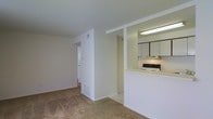 This single-floor residential space offers a clean, functional layout designed for comfortable everyday living. The open-concept living area is finished with wall-to-wall carpeting in a warm neutral tone, creating a cozy and welcoming atmosphere. The space is bathed in a light color palette throughout, with white walls and ceilings that enhance the sense of openness.

The kitchen is positioned adjacent to the living area and features a pass-through counter design that promotes connectivity between the two spaces. It is equipped with white cabinetry, overhead fluorescent lighting, and a range with an overhead ventilation hood. The cabinetry provides ample storage, while the countertop pass-through offers a practical surface for casual dining or serving.

A hallway leads from the main living area to additional rooms, suggesting a practical and well-organized floor plan. The corridor is well-lit and maintains the same bright, neutral aesthetic as the rest of the space. Doors along the hallway provide access to separate rooms, offering both privacy and versatility. Overall, the space presents a straightforward and efficient residential layout with a clean, move-in-ready appearance.