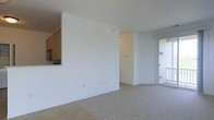 This single-floor space offers a clean, minimalist design characterized by bright white walls and neutral speckled flooring that runs throughout the main living area. The open layout provides a versatile living space that flows naturally from one area to the next. A pass-through counter connects the kitchen to the main living area, offering a practical and social design element. The kitchen, visible from the living space, features warm wood cabinetry and includes built-in appliances such as a microwave. A sliding glass door on one end of the space allows ample natural light to filter in, contributing to the overall bright and airy ambiance of the interior. The white walls and light-toned flooring enhance the sense of openness throughout. The layout is straightforward and functional, with smooth transitions between the kitchen and living areas. The space presents a blank canvas well-suited for a variety of interior arrangements and styles, with its neutral palette and clean architectural lines serving as a versatile foundation for personalization.