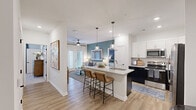 Located at 1215 Evolve Way in Greenville, NC, this thoughtfully designed 2-bedroom, 3-bathroom residence offers a modern, comfortable living experience across a single floor. The open-concept kitchen and living area serves as the heart of the home, featuring light wood-style flooring, a teal accent wall, and abundant natural light from large windows and French doors leading to a private balcony. The kitchen is outfitted with two-tone cabinetry—white uppers and dark lowers—a granite-topped island with pendant lighting and bar seating, a tile backsplash, and stainless steel appliances, including a refrigerator and dishwasher. A pantry adds convenient storage. The living area flows seamlessly from the kitchen and is topped with a ceiling fan for year-round comfort. Both bedrooms are carpeted and feature ceiling fans and generous closet space, with the primary bedroom showcasing vaulted ceilings and access to a walk-in closet with built-in wire shelving. The three bathrooms include a spa-like primary bath with a walk-in shower and dual granite-top vanity, and a secondary bath with a soaking tub and shower combo. A dedicated laundry room houses a full-size washer and dryer with overhead shelving, complementing a utility room, hallway, and additional closets throughout.