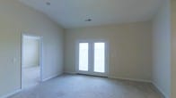 This single-floor space is designed with comfort and openness in mind. The main living area immediately draws attention with its vaulted ceilings, which lend a sense of generous vertical space and an airy feel. Neutral-toned walls complement the soft, plush carpeting underfoot, creating a warm and understated aesthetic throughout.

A standout feature of the living area is the set of white-framed French doors, which flood the room with abundant natural light and provide direct access to an outdoor balcony. The view through the doors hints at an exterior railing, suggesting an elevated position with outdoor enjoyment potential.

Visible HVAC venting in the ceiling indicates a climate-controlled environment, ensuring year-round comfort. The clean white baseboards and trim throughout provide a crisp, finished look that contrasts neatly with the beige walls. A doorway on the left side of the main room leads to an additional area of the space, indicating a functional and connected layout.

Overall, the space presents a clean, move-in ready environment with practical design choices, ample natural light, and architectural details that contribute to a bright and welcoming atmosphere.