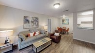 This single-story residence is thoughtfully laid out across multiple rooms, offering a functional and comfortable living arrangement. The living area serves as a welcoming focal point, featuring wood-look hardwood-style flooring, neutral walls, and a sliding glass door fitted with vertical blinds that allows natural light to filter through. The space flows directly into a dedicated dining area, which accommodates a round four-seat table and is finished with matching flooring and a textured ceiling.

The galley-style kitchen is efficiently designed with white cabinetry lining both sides, contrasting dark countertops, black appliances including a refrigerator and electric range, a stainless steel sink, and wood-look flooring underfoot. An adjoining utility area provides additional storage and cabinet space, with carpeted flooring transitioning toward the bedroom wing.

Multiple bedrooms are present throughout the unit. Each room is generously proportioned with plush carpeting, ceiling fans with integrated lighting, textured ceilings, and vertical blind-covered windows or sliding glass doors that provide outdoor views and natural light. The primary bedroom is notably spacious, offering room for a seating area alongside the bed. Throughout, the overall ambiance is clean, neutral, and move-in ready.