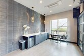 Located in Charlotte, NC, this thoughtfully designed rooftop amenity space offers a compelling blend of modern interior finishes and open-air outdoor living. The interior is anchored by a sleek wet bar area, where floor-to-ceiling textured slate-toned tile creates a dramatic accent wall. Slate-blue cabinetry with a crisp white countertop, brass fixtures, and a built-in beverage cooler complete the bar setup. Elegant glass pendant lights with gold hardware hang overhead, adding warmth to the sophisticated palette. Light porcelain tile flooring runs throughout, complementing the cool, contemporary aesthetic. The hallway makes a bold artistic statement with a vibrant, large-scale abstract mural that serves as a striking focal point amid neutral tile walls and clean-lined architecture. The expansive balcony is a true highlight of the space, featuring warm hardwood deck tiles laid in a herringbone pattern beneath a vivid teal-painted ceiling. A long communal bar-height table with stools invites gatherings, while a lounge area with outdoor seating and a wall-mounted television adds comfort. Structural cable railings and open sightlines frame sweeping panoramic views of the Charlotte skyline and surrounding greenery, making this an exceptional space for entertaining or relaxing.