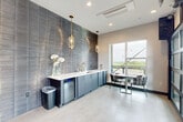 Located in Charlotte, NC, this thoughtfully designed rooftop amenity space offers a compelling blend of modern interior finishes and open-air outdoor living. The interior is anchored by a sleek wet bar area, where floor-to-ceiling textured slate-toned tile creates a dramatic accent wall. Slate-blue cabinetry with a crisp white countertop, brass fixtures, and a built-in beverage cooler complete the bar setup. Elegant glass pendant lights with gold hardware hang overhead, adding warmth to the sophisticated palette. Light porcelain tile flooring runs throughout, complementing the cool, contemporary aesthetic. The hallway makes a bold artistic statement with a vibrant, large-scale abstract mural that serves as a striking focal point amid neutral tile walls and clean-lined architecture. The expansive balcony is a true highlight of the space, featuring warm hardwood deck tiles laid in a herringbone pattern beneath a vivid teal-painted ceiling. A long communal bar-height table with stools invites gatherings, while a lounge area with outdoor seating and a wall-mounted television adds comfort. Structural cable railings and open sightlines frame sweeping panoramic views of the Charlotte skyline and surrounding greenery, making this an exceptional space for entertaining or relaxing.