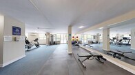 This fitness center offers a comprehensive, single-floor workout facility thoughtfully designed to accommodate a wide range of exercise routines. The space is divided into distinct functional zones, each catering to different training needs. The cardio area is lined with treadmills, an elliptical trainer, and a stationary bike, all positioned near large windows that invite natural light and views of surrounding greenery, creating an energizing atmosphere. A dedicated strength training section features a multi-function cable machine, a full dumbbell rack with a variety of weights neatly organized, and an adjustable workout bench. The free-form exercise zone is equipped with rubber foam flooring tiles, stability balls in assorted sizes, resistance bands, and exercise mats, providing ample space for stretching, core work, and functional training. Large wall-mounted mirrors run along one side of the room, visually expanding the space and allowing users to monitor their form. The neutral warm-toned walls and clean white ceiling contribute to a bright and motivating environment. A wall-mounted split air conditioning unit ensures comfortable temperatures year-round. A water cooler station near the entrance adds convenience. The space is well-lit with both natural and recessed lighting throughout.