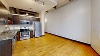 This fourth-floor unit at 101 E Sixth St in Winston-Salem, NC showcases an industrial-chic aesthetic rooted in its building's historic character. Rich hardwood floors run throughout, complemented by exposed ductwork, concrete columns, and high ceilings with track lighting. The open-concept living area flows into a well-appointed kitchen featuring dark wood cabinetry, granite countertops, and stainless steel appliances. A dramatic grid-pane factory window floods the space with natural light and offers urban views of the surrounding brick architecture. The bedroom includes a dedicated closet, and a hallway connects the layout efficiently.