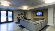 This single-floor space is designed as a stylish, contemporary lounge or entertainment area. The room centers around a generous light gray sectional sofa paired with dark upholstered ottomans, all arranged atop a layered area rug that anchors the seating zone. A standout feature is the rustic wood-paneled accent wall, which contrasts warmly against the otherwise neutral gray walls and provides a natural, textured backdrop for the wall-mounted flat-screen television below. Built-in cabinetry flanks the TV wall, offering practical storage while maintaining a clean aesthetic. On the opposing wall, an eclectic arrangement of floating box shelves adds visual interest and display space. The ceiling is smooth and finished with a flush-mount drum pendant light that provides warm, even illumination throughout the room. The flooring appears to be a polished concrete or tile, contributing to the space's modern, urban character. A glass-paneled door visible on the left side of the room introduces natural light and connects the interior to an adjacent area or exterior. Overall, the space blends industrial and contemporary design elements for a welcoming yet refined atmosphere.