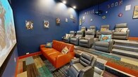 Located at 7700 Lincoln Ave NE in Rio Rancho, New Mexico, this dedicated movie room delivers an immersive, comic book-inspired home theater experience. The space is defined by deep royal blue walls and a dark ceiling, creating a classic cinema atmosphere enhanced by recessed overhead lighting. A large projection screen anchors the front wall, paired with a ceiling-mounted projector for a true theatrical viewing setup. Surround sound speakers are mounted at strategic points along the walls to complete the audiovisual experience. Seating is arranged in a tiered layout, with multiple rows of gray leather reclining chairs elevated on stepped platforms trimmed in bold orange — a nod to traditional cinema-style stadium seating. Complementing these are a vibrant orange leather sofa and additional gray leather recliners positioned at floor level near the screen. The flooring features a colorful, multi-toned patterned carpet that adds energy and visual interest throughout the room. The walls are adorned with framed comic book artwork, hexagonal decorative accents, and bold lettering spelling out "MOVIE ROOM," reinforcing the playful superhero theme. Comic book-themed throw pillows with phrases like "ZAP!" and "POW!" add fun, character-driven detail to each seat. A single entry door with a film reel motif and a staircase with an orange handrail complete this thoughtfully designed entertainment space.