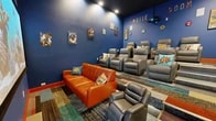 Located at 7700 Lincoln Ave NE in Rio Rancho, New Mexico, this dedicated movie room delivers an immersive, comic book-inspired home theater experience. The space is defined by deep royal blue walls and a dark ceiling, creating a classic cinema atmosphere enhanced by recessed overhead lighting. A large projection screen anchors the front wall, paired with a ceiling-mounted projector for a true theatrical viewing setup. Surround sound speakers are mounted at strategic points along the walls to complete the audiovisual experience. Seating is arranged in a tiered layout, with multiple rows of gray leather reclining chairs elevated on stepped platforms trimmed in bold orange — a nod to traditional cinema-style stadium seating. Complementing these are a vibrant orange leather sofa and additional gray leather recliners positioned at floor level near the screen. The flooring features a colorful, multi-toned patterned carpet that adds energy and visual interest throughout the room. The walls are adorned with framed comic book artwork, hexagonal decorative accents, and bold lettering spelling out "MOVIE ROOM," reinforcing the playful superhero theme. Comic book-themed throw pillows with phrases like "ZAP!" and "POW!" add fun, character-driven detail to each seat. A single entry door with a film reel motif and a staircase with an orange handrail complete this thoughtfully designed entertainment space.