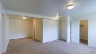 This single-floor space presents a generously sized room finished with neutral-toned wall-to-wall carpeting that contributes to a warm, quiet atmosphere. The white walls are clean and well-maintained, offering a bright, move-in-ready aesthetic. Architectural interest is provided by a structural beam that subtly divides the ceiling plane, adding modest dimensionality to the layout. Multiple flush-mount ceiling light fixtures are evenly distributed throughout, ensuring consistent illumination across the entire space. Several white-paneled doors line the far wall, suggesting access to closets or adjoining rooms, which adds to the overall storage and functional potential of the space. A partial wall segment near the right side of the room introduces a secondary nook or passage area, hinting at a thoughtful floor plan with defined zones. The smooth, white ceiling complements the light walls to create an open, airy feel. Overall, the space presents a clean, neutral canvas well-suited for a variety of furnishing arrangements and uses.