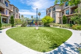 Located at 14041 Locust St in Westminster, California, this single-level communal outdoor space is set within a Mediterranean-inspired residential complex characterized by warm stucco exteriors, red-tiled rooflines, and balconied upper floors that frame the grounds on multiple sides. The central feature of the space is a generously sized, well-kept lawn area that serves as an open gathering space, providing a bright and inviting atmosphere under Southern California's abundant sunshine.

A covered picnic and seating area anchors one side of the lawn, offering shaded outdoor dining beneath a modern pergola-style canopy with multiple picnic tables. Adjacent to this structure, a colorful children's playground with slides and play equipment provides a dedicated recreational area for younger residents. The playground is set on a rubberized safety surface, ensuring a secure environment for children.

Curving aggregate pathways wind throughout the grounds, connecting various areas of the complex and lined with thoughtfully arranged drought-tolerant plants, ornamental grasses, shrubs, and mature trees. These landscaped borders add lush greenery and visual depth while complementing the warm tones of the building exteriors. The overall ambiance is open, family-friendly, and well-maintained, making it an appealing shared outdoor environment.