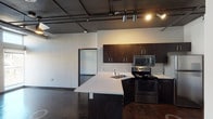 This single-floor unit showcases a compelling industrial-loft aesthetic defined by polished concrete floors, exposed ceiling ductwork, and dark ceiling panels that run throughout the space. The open-concept kitchen is a functional centerpiece, featuring dark espresso cabinetry, light quartz countertops, a peninsula island with an integrated sink, and a full suite of stainless steel appliances including a range, over-the-range microwave, and refrigerator. Track lighting along the exposed ceiling structure illuminates the kitchen and living areas effectively.

The living and sleeping areas are generously proportioned with high ceilings and large floor-to-ceiling windows that draw in substantial natural light while offering views of the surrounding streetscape. Industrial-style pendant lights hang throughout, complementing the raw architectural character of the space. A ceiling fan provides additional comfort in one of the rooms.

The bathroom continues the unit's design language, featuring polished concrete flooring, a clean white vanity with a wide countertop, a full-size soaking tub with shower, and simple chrome fixtures. Built-in closet organizers are visible in adjacent spaces, providing practical storage. The overall ambiance is modern and urban, balancing raw industrial elements with clean, functional finishes.