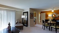 This single-floor residence offers a thoughtfully arranged open-concept layout that seamlessly connects the living, dining, and kitchen areas. The living space is finished with plush carpeting and neutral-toned walls, creating a warm and inviting atmosphere. A large floor-length mirror and a comfortable upholstered armchair with ottoman add both style and functionality to the room. A floor lamp provides warm ambient lighting, complementing the natural light that filters in through vertical blinds on a nearby window. The dining area transitions naturally from the living space, featuring a dark-toned table and chairs set against a rich, warm accent wall that adds a bold focal point to the open floor plan. The kitchen is visible through an open passageway and is equipped with wooden cabinetry and standard appliances. A notable convenience of this unit is the in-unit laundry area, tucked neatly alongside the kitchen, housing a front-loading washing machine. A white paneled door leads to an additional room, offering further privacy and versatility. The overall design balances practical living with a cozy, residential aesthetic throughout.