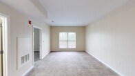This single-floor unit presents a practical and well-organized layout, making efficient use of its interior space. The living area is finished with neutral-toned carpet and warm beige walls, creating a calm, understated ambiance. A centrally placed window allows natural light to fill the room, offering views of the surrounding greenery outside.

The kitchen is a functional highlight of the unit, featuring rich medium-toned wood cabinetry with ample upper and lower storage. Black appliances — including an electric range, refrigerator, and built-in range hood — complement the cabinetry. A peninsula with a built-in sink and light countertops provides additional workspace and separates the kitchen from the adjacent area. Light-toned tile flooring runs throughout the kitchen, offering easy maintenance.

The bedrooms continue the neutral carpet and beige wall palette, each featuring windows that admit natural light. The rooms offer versatile, open layouts suitable for various furniture configurations.

The bathroom includes a single-sink vanity with a wood cabinet base and white countertop, a full tub and shower combination with chrome fixtures, and light speckled tile flooring. The overall aesthetic of the unit is consistent and clean throughout.