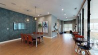 Located at 2599 Evans Rd in Morrisville, NC, this single-level amenity space is thoughtfully designed to support both collaborative work and casual gathering. The layout flows seamlessly from a formal meeting area into an open lounge and co-working zone, all unified by warm wood-look tile flooring and a neutral gray color palette.

The conference area serves as a focal point, anchored by a large rectangular table with rich leather-upholstered chairs. A wall-mounted flat-screen display and a pair of geometric pendant lights overhead lend a polished, professional feel. A dramatic deep teal textured accent wall and a set of oversized abstract art panels add bold visual interest.

Transitioning into the open lounge, floor-to-ceiling glass panel partitions with sleek black metal frames define the space while preserving sightlines and allowing abundant natural light to pour in from large exterior windows. Exposed white brick columns add an industrial yet refined architectural detail. Bistro-style seating with sculptural chairs provides casual gathering spots, while a dedicated workstation area along one wall accommodates individual productivity needs. Decorative shelving, curated accessories, and framed artwork throughout contribute to a cohesive, gallery-like atmosphere. The space balances functionality with design sophistication, making it well-suited for both focused work and social interaction.